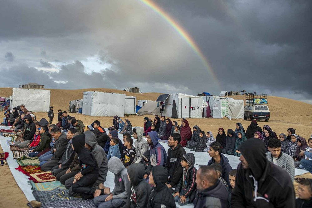 Palestinian Muslims in Gaza perform the Eid al-Fitr prayer in an open space, with destruction in the background and a rainbow overhead.