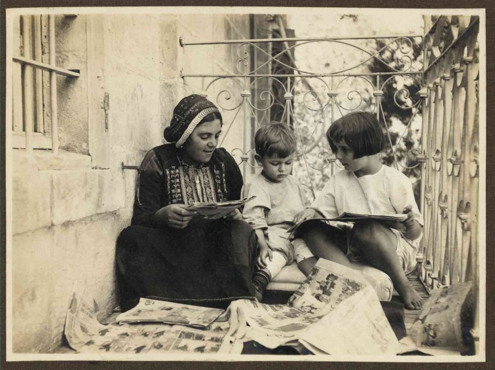 Woman dressed in traditional Palestinian embroidery reads newspaper cartoons to children