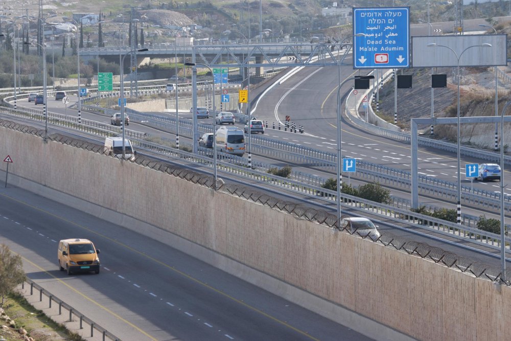 A Palestinian taxi drives on one side of the Separation Wall, while Israeli vehicles drive on the other side