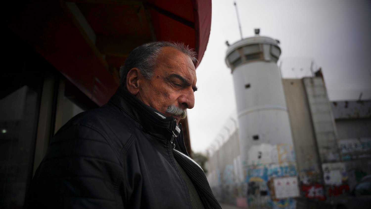 Abu Alzoz Nassar, owner of al-Karawan Restaurant, is overshadowed by a Separation Wall guard tower, 2021
