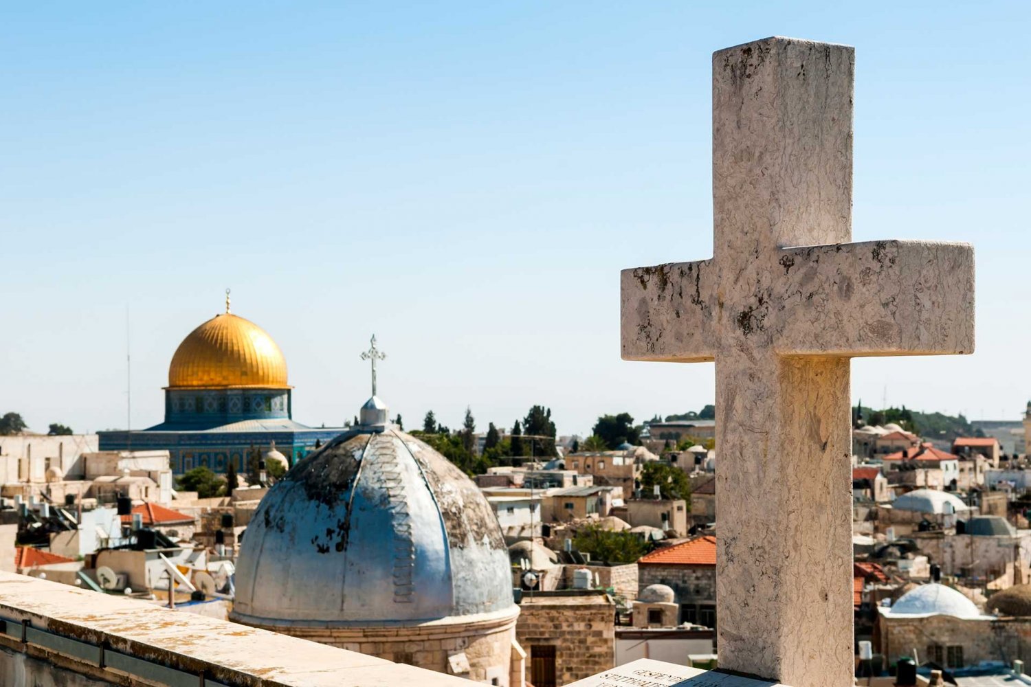 A cross atop a building in the Old City of Jerusalem. In the background are two domes, one a church with a cross and the other the Dome of the Rock with a crescent.